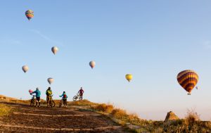 Group of friends with mountain bikes on a trip in the region of Cappadocia, Turkey. In an early morning, as they follow their route, hot air balloons start rising up above them.